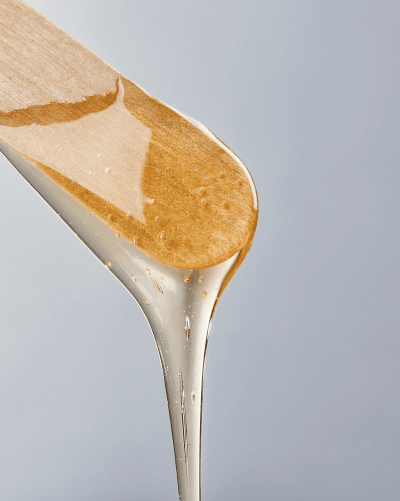 Honey dripping from a wooden honey dipper against a light gray background.